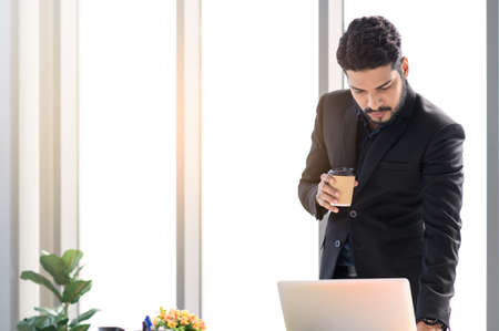 Thoughtful businessman wearing black suit and holding paper coffee cup, checking documents in laptop computer stand at office desk. Big window in the background. Pensive manager reading reports or checking dataの写真素材