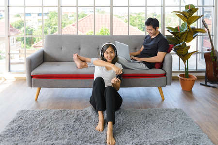 Lovely Asian couple in living room at home. young woman with headphone sit on floor playing video game, man lying on couch working with laptop computer in background at home. Love, happiness, WFH, work from home conceptの写真素材