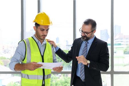 Construction engineer in vest and helmet discussing with construction manager, standing on construction siteの写真素材