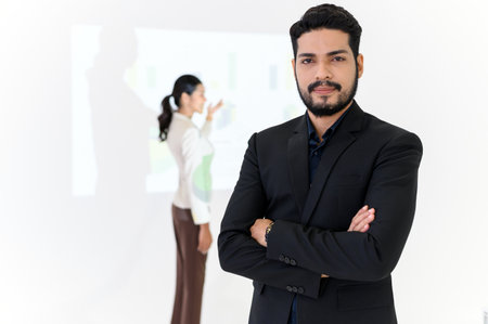 Confident bearded businessman smiling while standing and arms crossed, Businesswoman gives presentation on background, looking at the cameraの写真素材