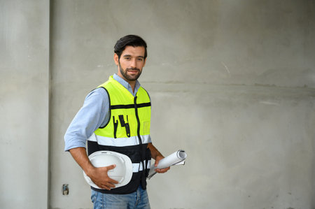 Portrait ofÂ young attractive construction man smiling bearded in vest and holding white helmet...