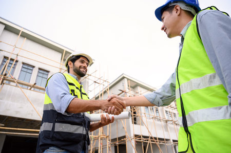 Construction engineer and architect with blueprint shaking hands while standing on construction...