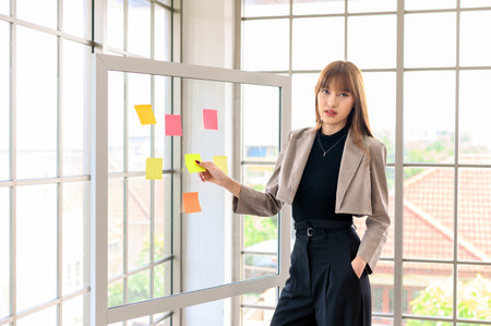 young modern businesswoman in casual fashion suit looking at the camera while using sticky notes for strategy ideas on glass board in board room at the office. Business project planningの写真素材