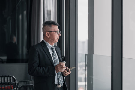 Thoughtful, mature businessman holding coffee paper cup and smartphone while standing and looking outside the windowの写真素材