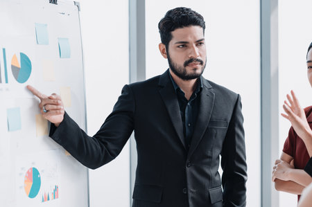 Confident businessman giving a presentation with charts and graphs on a whiteboard, explaining data analysis, strategy, and business growth to colleagues in a modern officeの写真素材