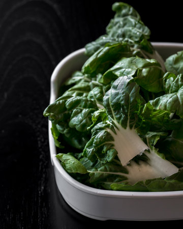 A bowl of green spinach leaves. The bowl is white and the spinach is fresh and green. The bowl is placed on a black surfaceの写真素材
