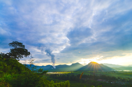 Smoke billows from a distant at sunrise, casting a dramatic light over a lush, green landscape with rolling hills andÂ aÂ vibrantÂ sky.の写真素材