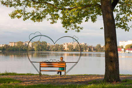 Young woman is sitting and waiting on a bench with big heart on itの写真素材