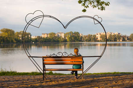 Young woman is sitting and waiting on a bench with big heart on itの写真素材