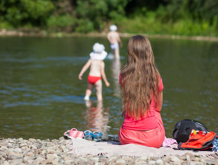 Young mother looking after her children on the bank of the river on summer sunny dayの写真素材
