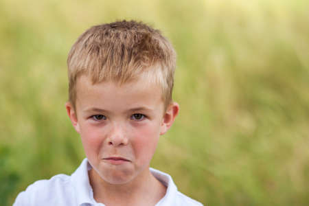 Portrait of a little displeased and dissatisfied boy with golden blonde straw hair in sunny summer day on green blurred backgroundの写真素材