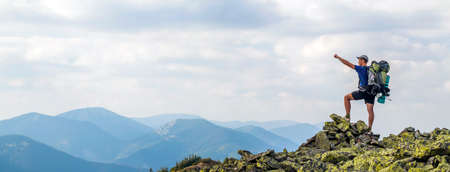 Man on peak of mountain. Emotional scene. Young man with backpack standing with raised hands on top of a mountain and enjoying mountain view. Hiker on the mountain top. Sport and active life concept.の写真素材