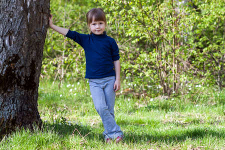 Lovely smiling little girl standing near big tree on green grass on a sunny dayの写真素材