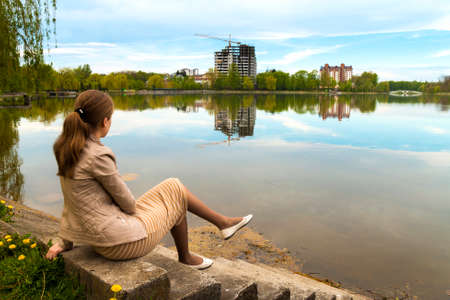 Beautiful young woman sitting on the bank of big lake with modern city skyline of buildings under constructionの写真素材