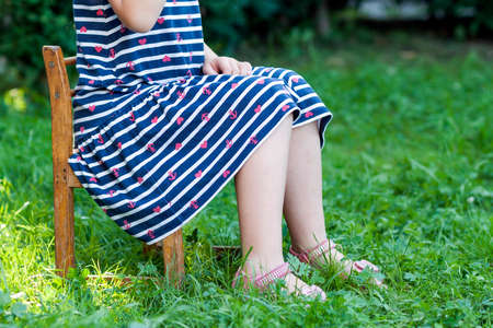 Legs and feet of little girl in dress sitting on a chair on green grassの写真素材