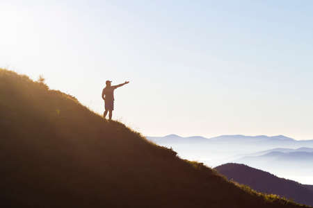 Man on peak of mountain. Emotional scene. Young man with backpack standing with raised hands on top of a mountain and enjoying mountain view. Hiker on the mountain top. Sport and active life concept.の写真素材
