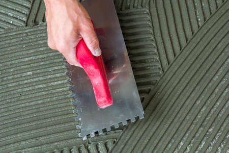 Close-up detail of floor tiles installation. Home improvement, renovation. Workers hand with notched float for tile. Ceramic tile floor adhesive, mortar.の写真素材