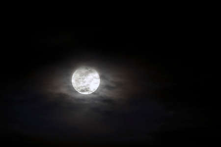 Big silver glowing Moon close-up on dark blue sky with scattered clouds.