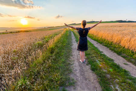 Young girl standing on dirt gravel road in wheat field at sunset raising handsの写真素材