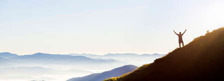 Panorama of young successful man hiker silhouette with open arms on morning foggy mountain peak.の写真素材