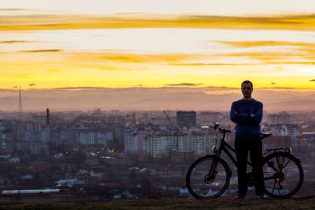 Dark silhouette of man standing near a sports bicycle with the night city view behind.