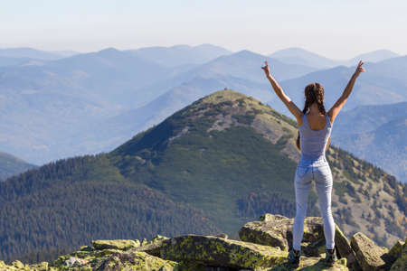 Silhouette of a young happy beautiful slim girl with long braids standing on big rocks in mountains lifting hands, meeting a raising sun. Tourism and traveling.の写真素材