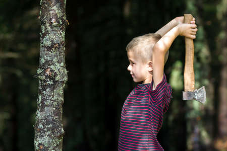 Small boy with heavy old iron axe cutting tree trump in forest on summer day. Outdoor activities and...