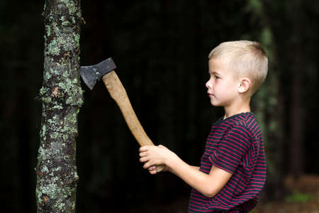 Small boy with heavy old iron axe cutting tree trump in forest on summer day. Outdoor activities and physical labor.の写真素材