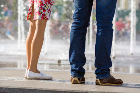 Close up of two pairs of legs, girl in short dress with floral pattern and white shoes and man in blue jeans and brown shoes,standing together on blurred colorful background.の写真素材