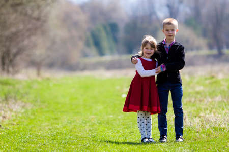 Portrait of beautiful children in new clothes standing together outdoors on bright blurred background, older brother embracing protectively sister. Happy friendship and family relations concept.の写真素材