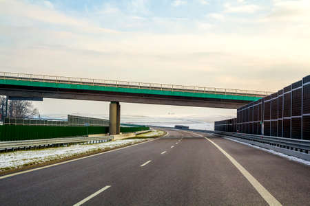 Modern wide smooth empty asphalt highway stretching to horizon under high bridge turning right past...