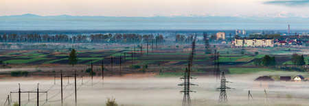 Beautiful panorama of long electric high voltage power line rows stretching to horizon through spring fields and trees under dense morning fog on distant Carpathian mountains range background.の写真素材