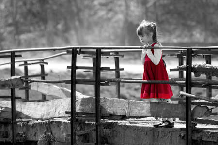 Black and white picture of pretty blond child girl in red dress standing alone on old bridge leaning on wooden railings looking down on warm sunny day. Children curiosity and fantasies concept.の写真素材