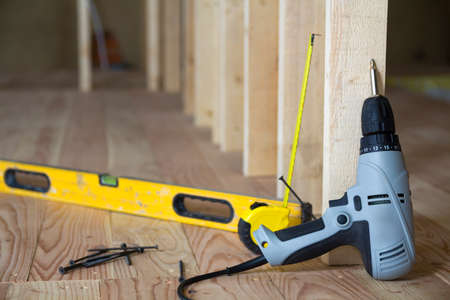 Close-up of professional tools: electrical screwdriver, level and measuring tape on background of wooden frame for future wall in attic room under reconstruction. Renovation and improvement concept.の写真素材