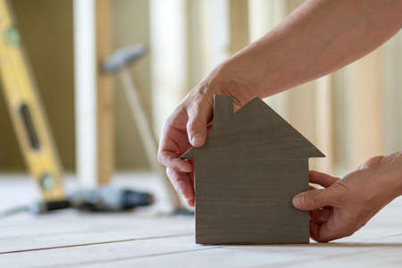 Close-up of muscular male hands holding small brown wooden model house on blurred background of...