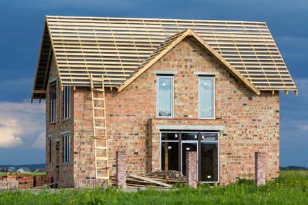 Two-story residential house with plastic windows, wide door and wooden frame for roof under construction. High wooden ladder at brick walls and brick columns for fence on dark blue sky background.の写真素材