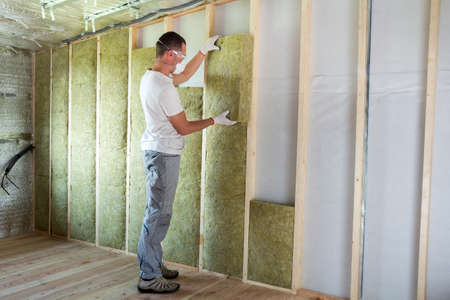 Worker in protective goggles and respirator insulating rock wool insulation in wooden frame for future house walls for cold barrier. Comfortable warm home, economy, construction and renovation conceptの写真素材