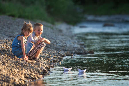Two smiling blond children, boy and girl playing with white paper boats on river bank on bright summer blurred blue background. Joys and games of happy childhood and outdoors activities concept.の写真素材