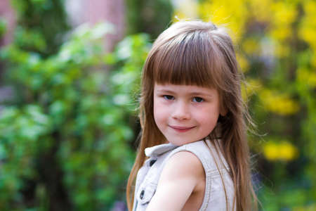 Portrait of a pretty little long-haired blond preschool girl in sleeveless white dress smiling shyly...
