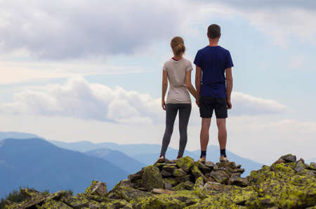Back view of young tourist couple, athletic man and slim girl stand holding hand on rocky mountain top enjoying breathtaking summer mountain panorama. Tourism, traveling and healthy lifestyle concept.の写真素材