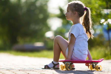 Profile portrait of pretty small long-haired blond girl in white clothing sitting on skateboard on bright summer day on blurred bokeh background. Children activities and active lifestyle concept.の写真素材