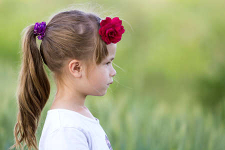 Profile portrait of cute small serious girl in white T-shirt with long blond pony-tail and red rose in hair on blurred bright green copy space background. Beauty of childhood concept.の写真素材
