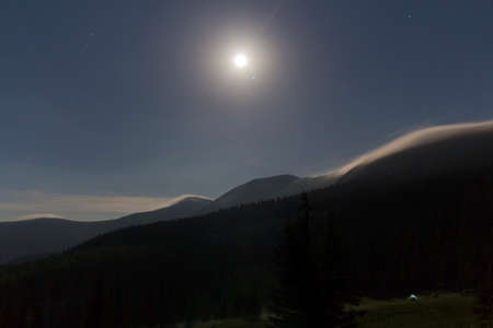 Night view of bright moon shining in dark blue sky over mountains covered with evergreen forest and...