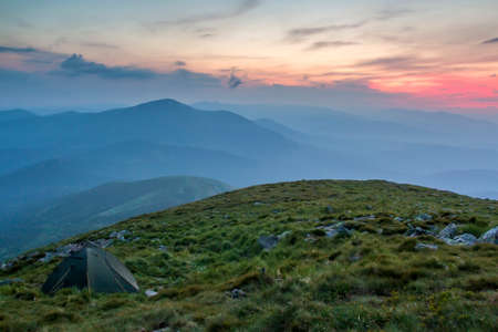 Summer camping in mountains at dawn. Tourist tent on round grassy hill on distant misty blue...