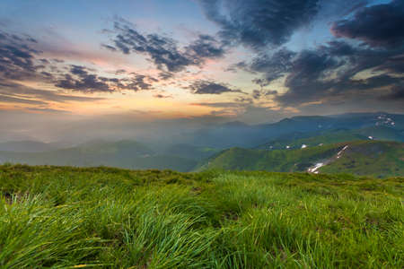 Wide summer mountain view at sunrise. Glowing orange sun raising in blue cloudy sky over green...
