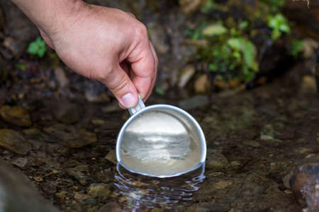 Close-up of man hand with tourist aluminum cup scooping clean fresh sweet water from natural...