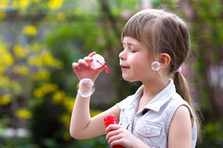 Profile of pretty small preschool blond girl with funny serious expression blowing colorful transparent soap bubbles outdoors on blurred green summer background. Joy of careless childhood concept.の写真素材