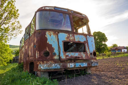 Close-up of old forsaken passenger bus with broken windows rusting in high green weedy grass on edge of plowed brown field on bright spring day under blue morning sky on small peasant huts background.の写真素材