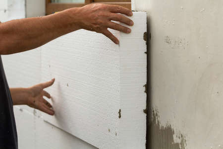 Worker hands installing white rigid polyurethane foam sheet on plastered brick wall. Modern...