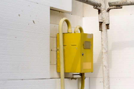 Close-up of yellow gas meter box with connecting pipes hanging on the corner of house wall insulated...